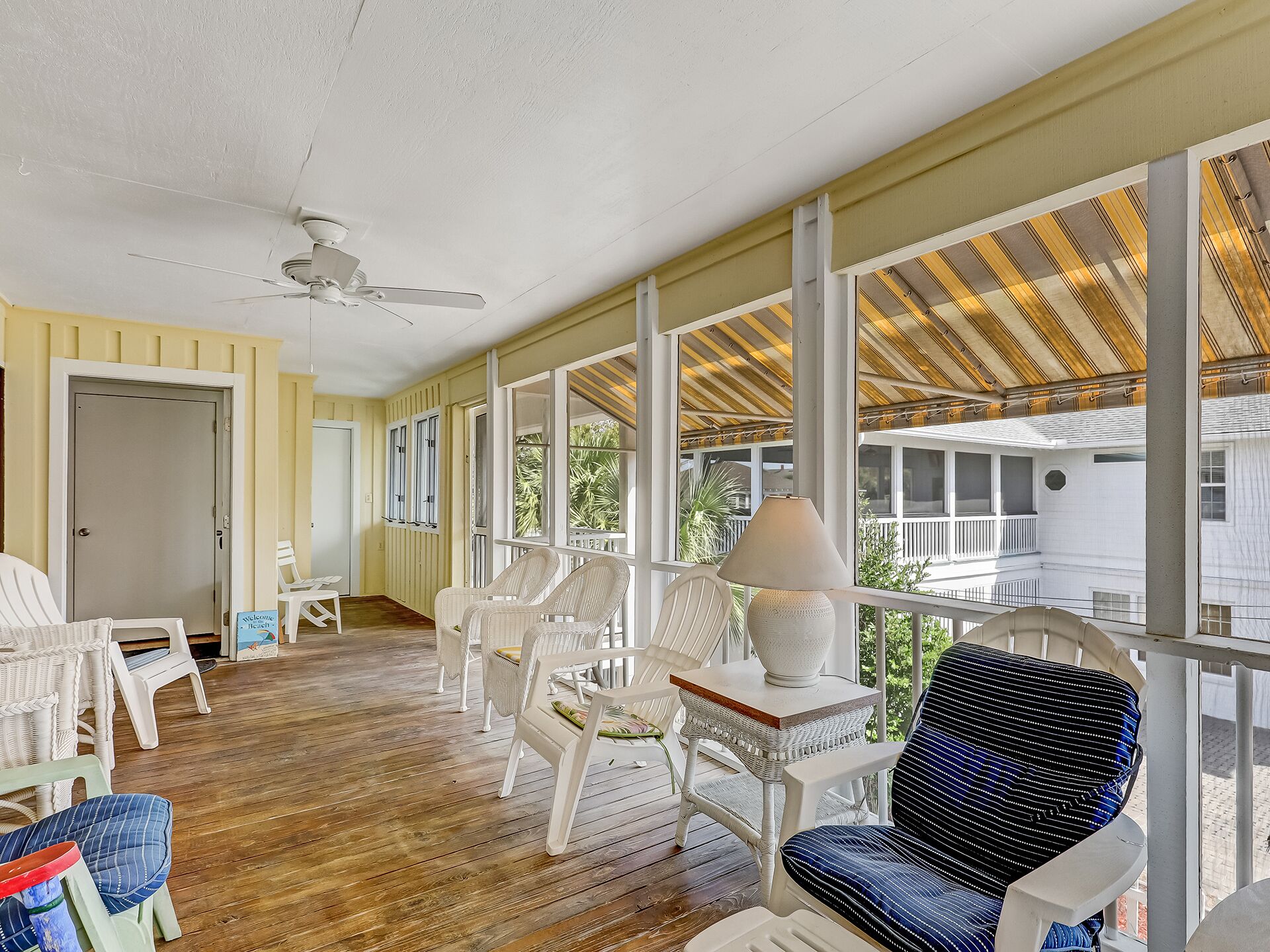 A covered porch with wicker chairs, a table with a lamp, and a view of a house.