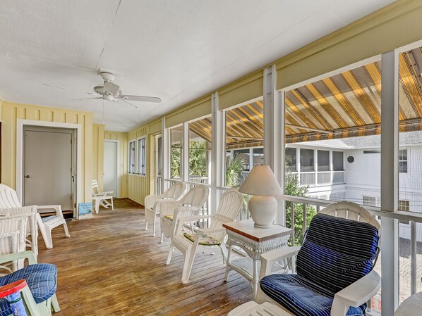A covered porch with wicker chairs, a table with a lamp, and a view of a house.