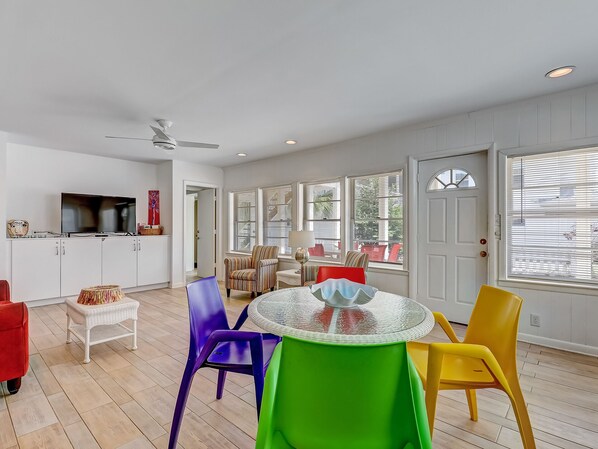 A spacious living area with a round glass table, colorful chairs, a television, and a door leading to another room. - Family Beach Getaway, Heated Pool Access (Tybee Island)