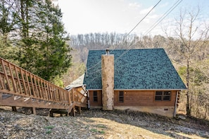 A log cabin with a stone chimney, wooden deck, and a view of the surrounding forest.