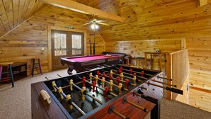 Une pièce au plancher de bois, équipée d’un baby-foot, d’une table de billard et d’un ventilateur de plafond.