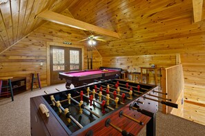 A wooden-floored room with a foosball table, pool table, and a ceiling fan.