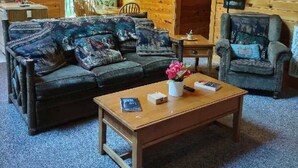 L’intérieur d’un chalet confortable, avec des murs en bois, un escalier en bois, un ensemble de canapés aux couleurs sombres, une petite table ornée d’un vase de fleurs et un fauteuil avec un coussin.