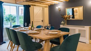 Ein Esszimmer mit einem Holztisch, gedeckt für sechs Personen, umgeben von grünen Stühlen. Eine weiße Tür und ein Fenster mit Vorhängen sind sichtbar, und ein Spiegel hängt an der Wand.
