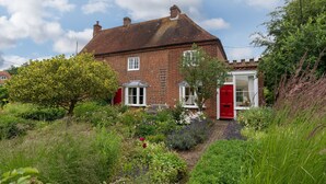Une maison en briques rouges, avec une porte et des fenêtres blanches, entourée d’une végétation luxuriante et d’un jardin.