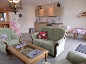 Ein Wohnzimmer mit zwei grünen Sesseln, einem Holztisch und einer Küche im Hintergrund.