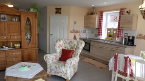 Een gezellige keuken met een fauteuil met bloemenprint, houten keukenkastjes en een eethoek met een tafel en stoelen.