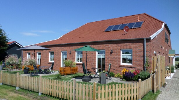 Een gebouw van baksteen met een rode dakbedekking, zonnepanelen, een houten omheining en terrasmeubels.