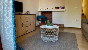 A living room with a wooden cabinet, a blue chair, a round white table with a geometric design, and a fireplace with decorative items.