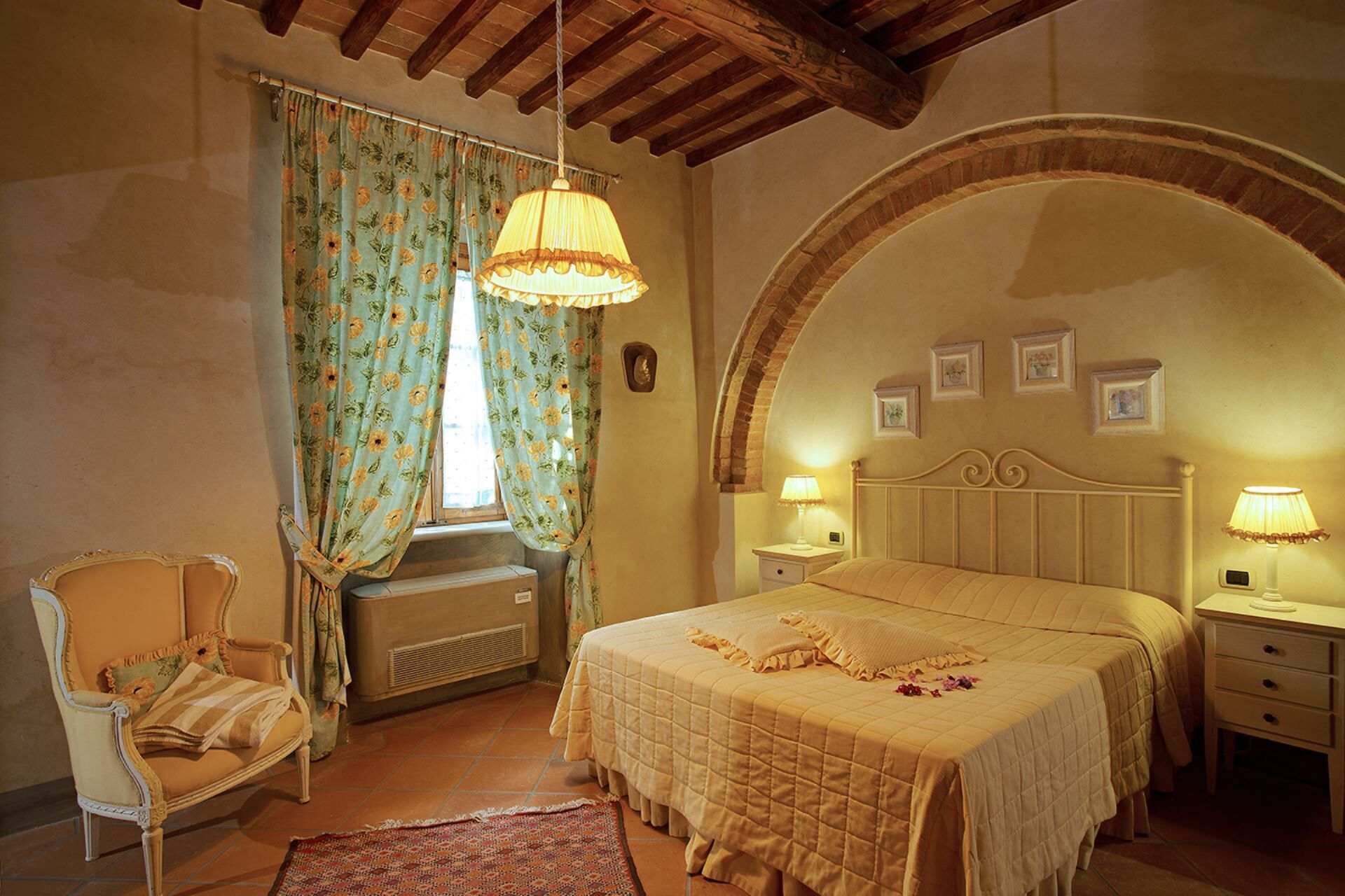 Una camera da letto con un letto, una sedia, una finestra con tende, un termosifone e un muro decorato con quadri.