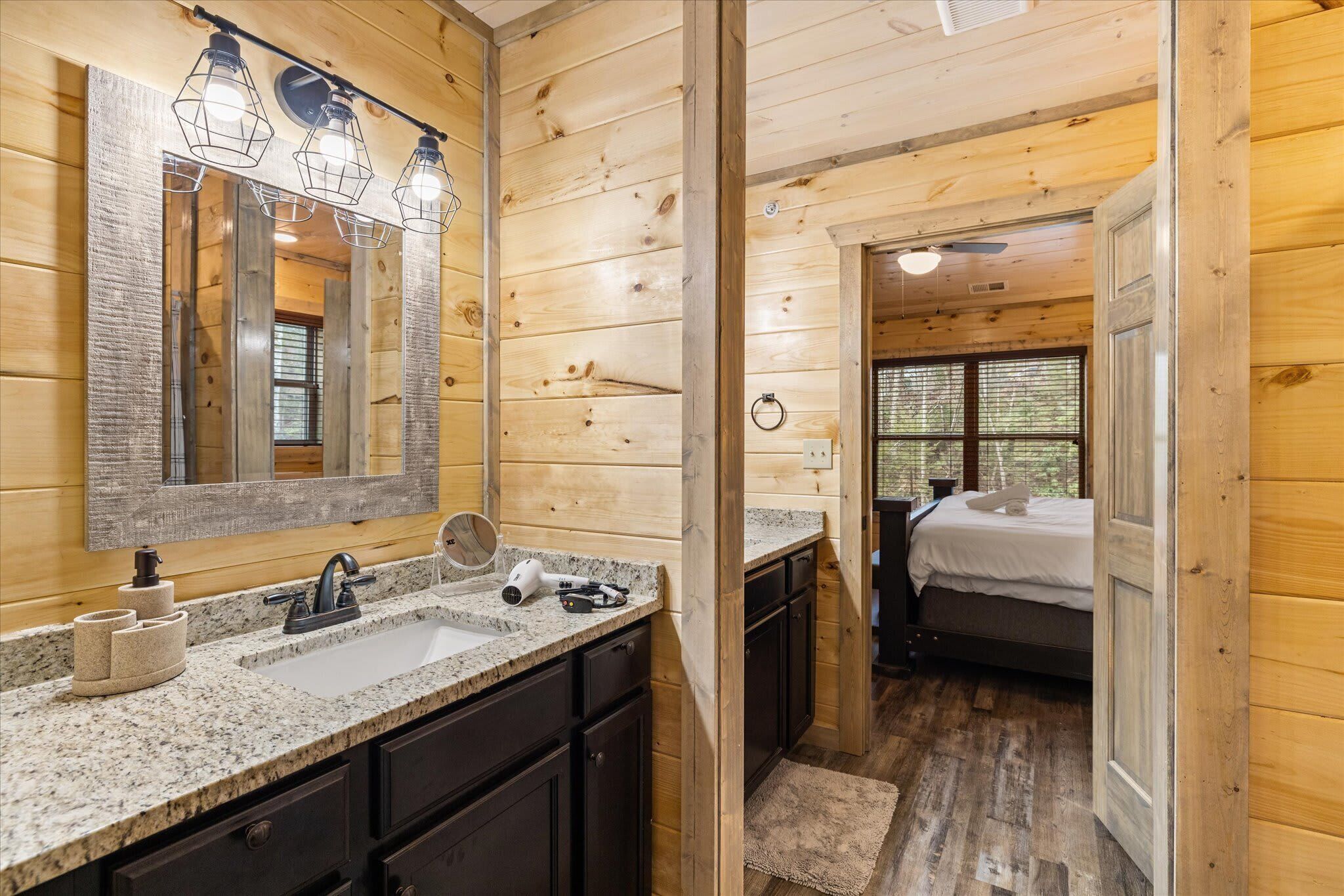 L’intérieur d’un chalet en bois comprend une salle de bain avec un lavabo doté d’un plan de travail en granit, un grand miroir et un sèche-cheveux. La chambre à coucher est équipée d’un lit avec une couette blanche et d’une fenêtre avec des stores.