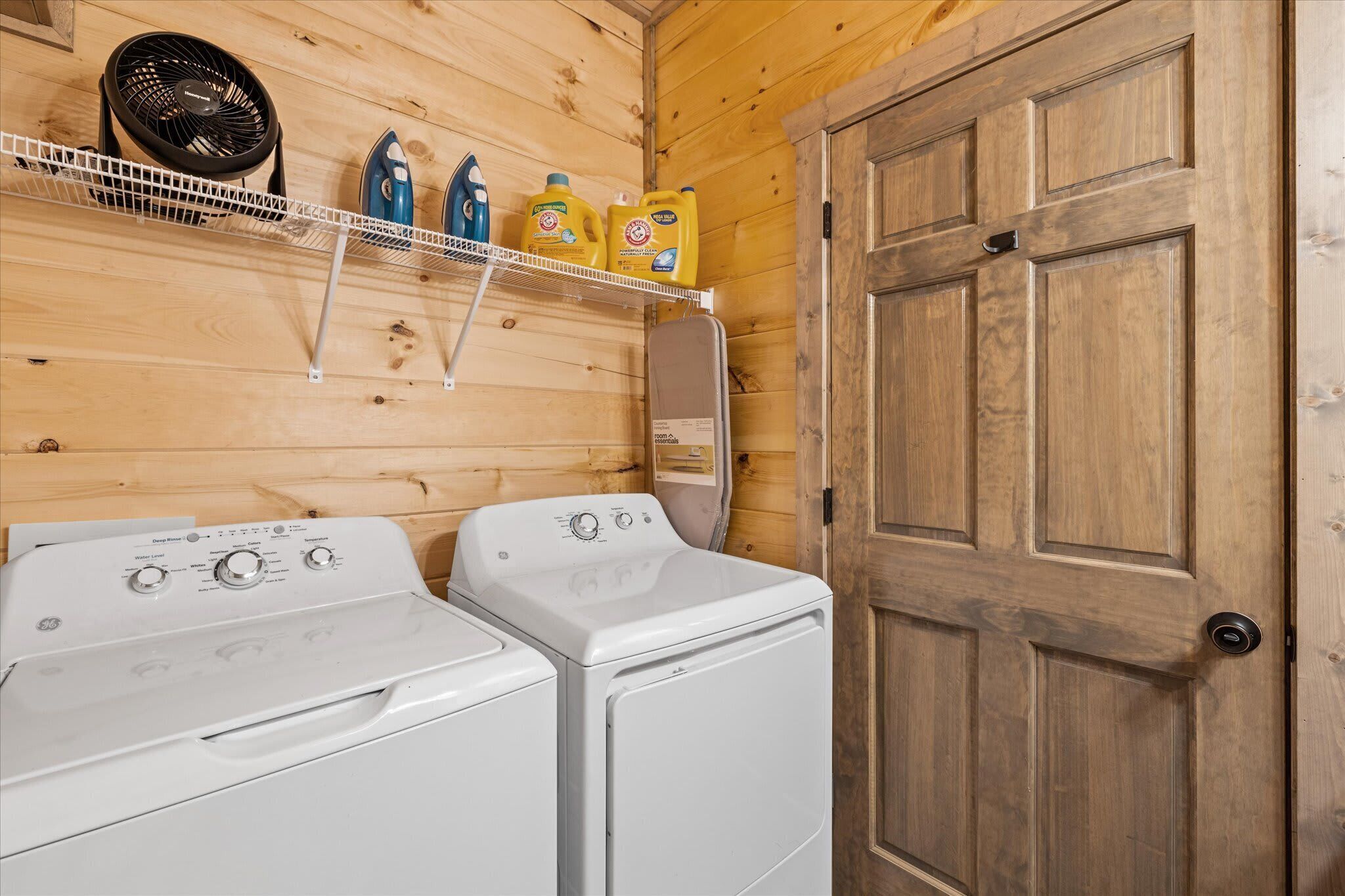 A laundry area with two white washing machines, a fan, and detergent on a shelf.