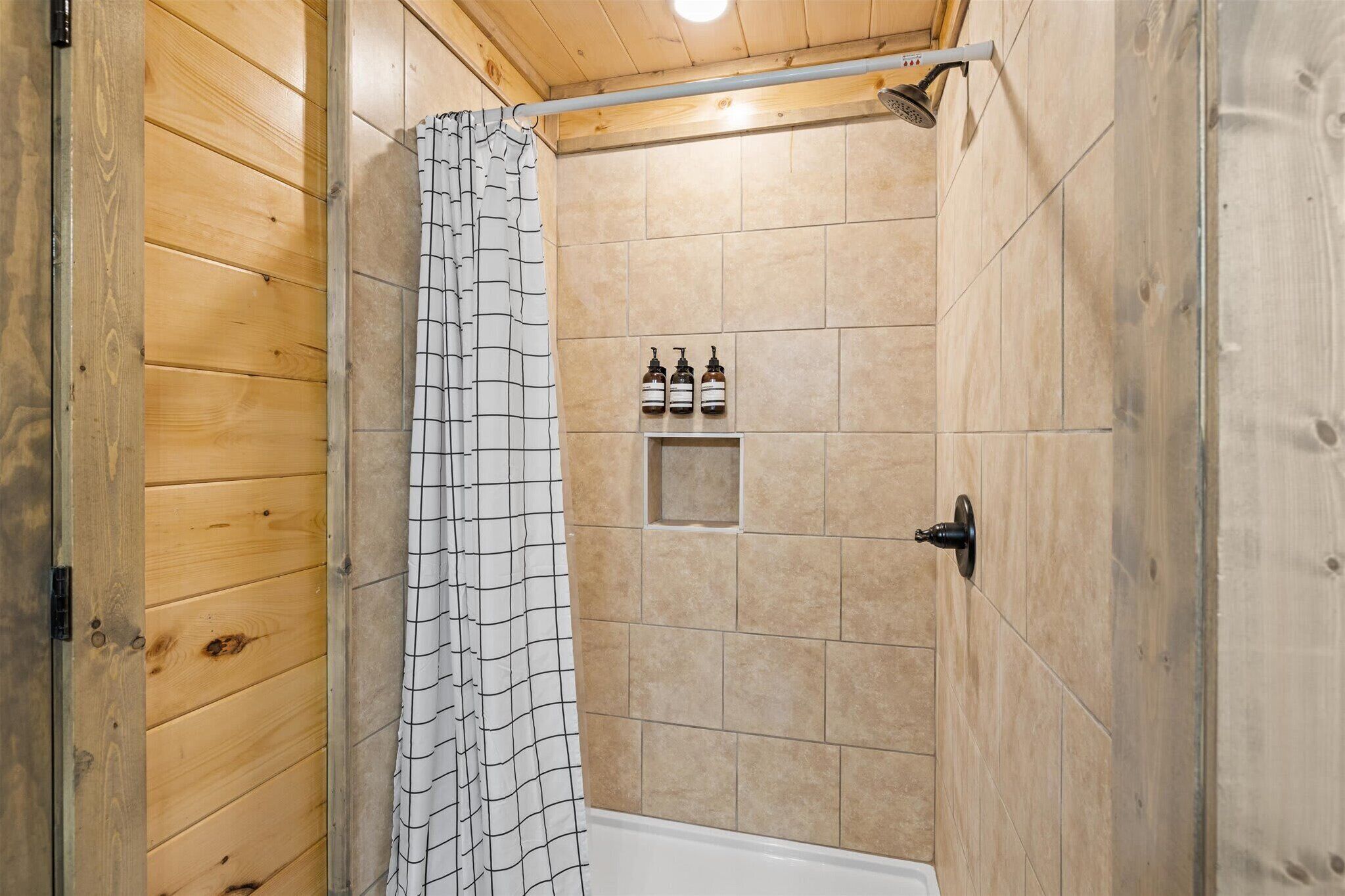 A wooden-paneled shower room with a tiled wall, a showerhead, and three bottles of shampoo on a shelf.