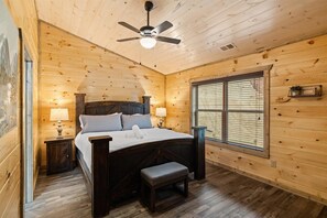 A wooden cabin bedroom with a bed, nightstands, a bench, a ceiling fan, and a window.