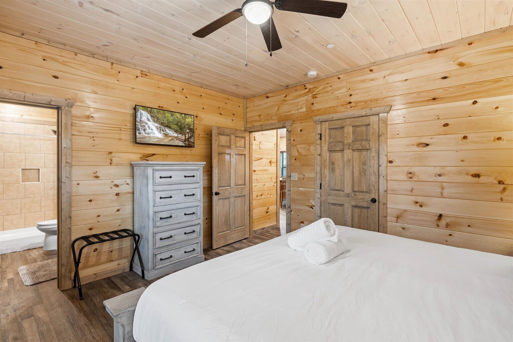 A wooden cabin bedroom with a bed, a dresser, a ceiling fan, and a painting on the wall.