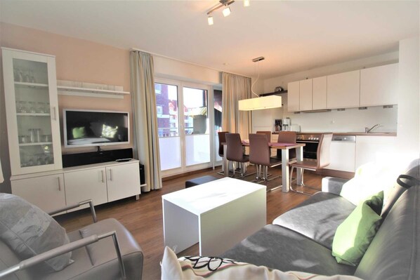 A modern living room with a flat-screen TV, a dining area with chairs, a kitchen with cabinets, and a large window.