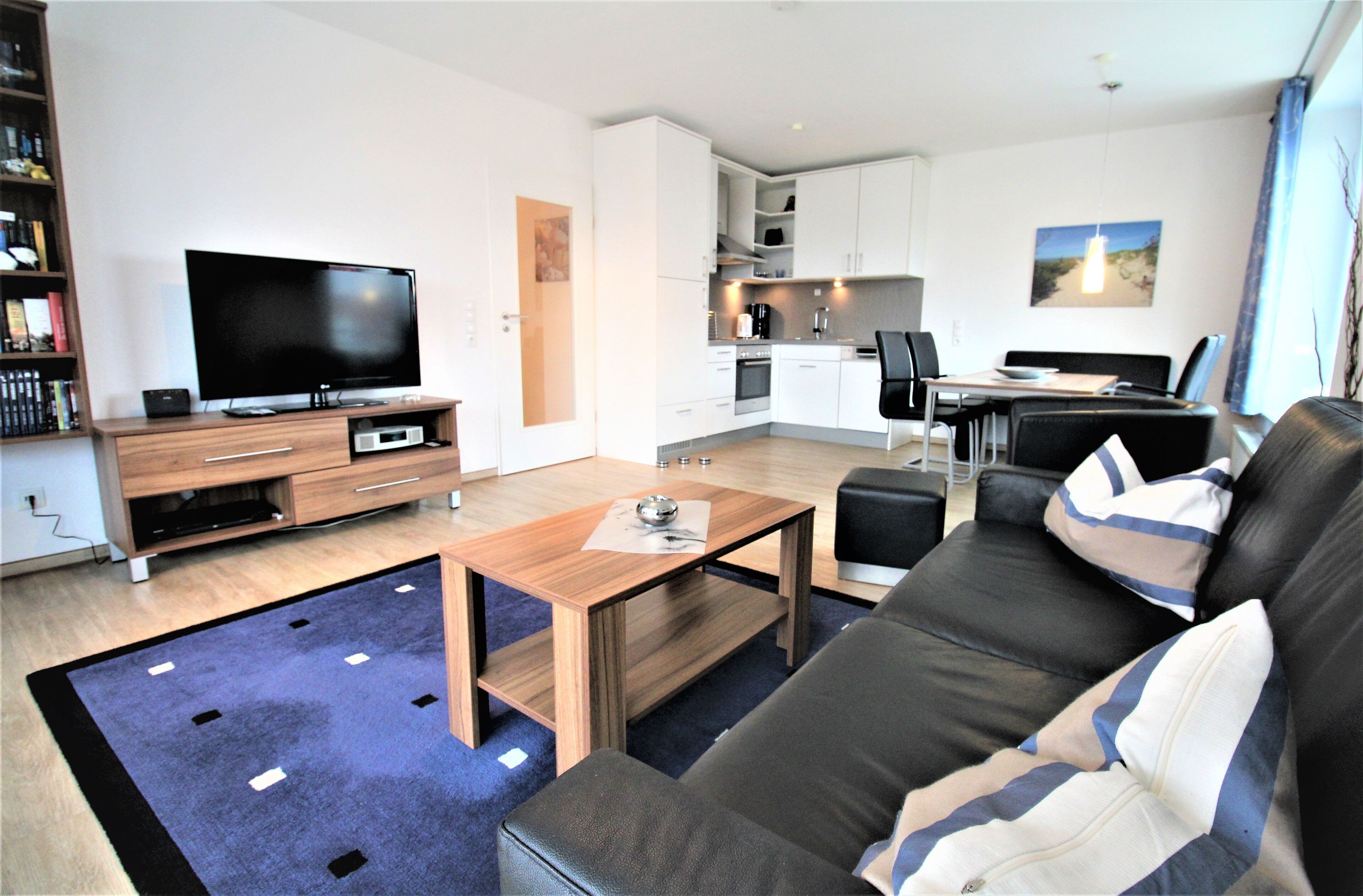 A modern living room with a flat-screen TV, a wooden coffee table, a black leather sofa, and a dining area with a table and chairs.