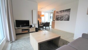 A modern living room with a flat-screen TV, a coffee table, a sofa, and a dining area with chairs and a table.