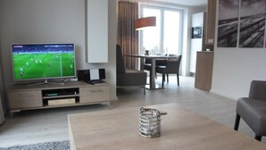 A modern living room with a flat-screen TV, a coffee table, a dining area with a table and chairs, and a large window with curtains.
