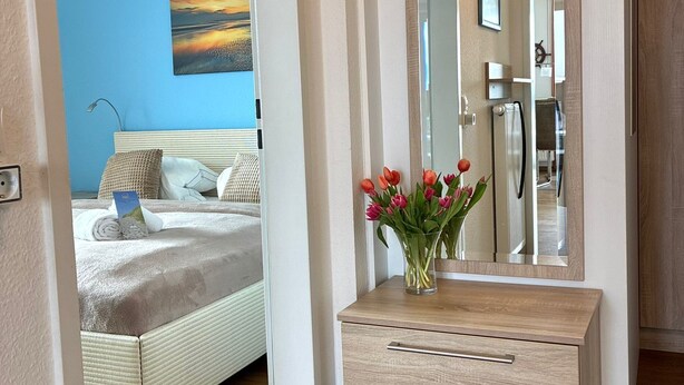 Un dormitorio con cama, una mesita de noche con un florero, un espejo grande y una pared azul.