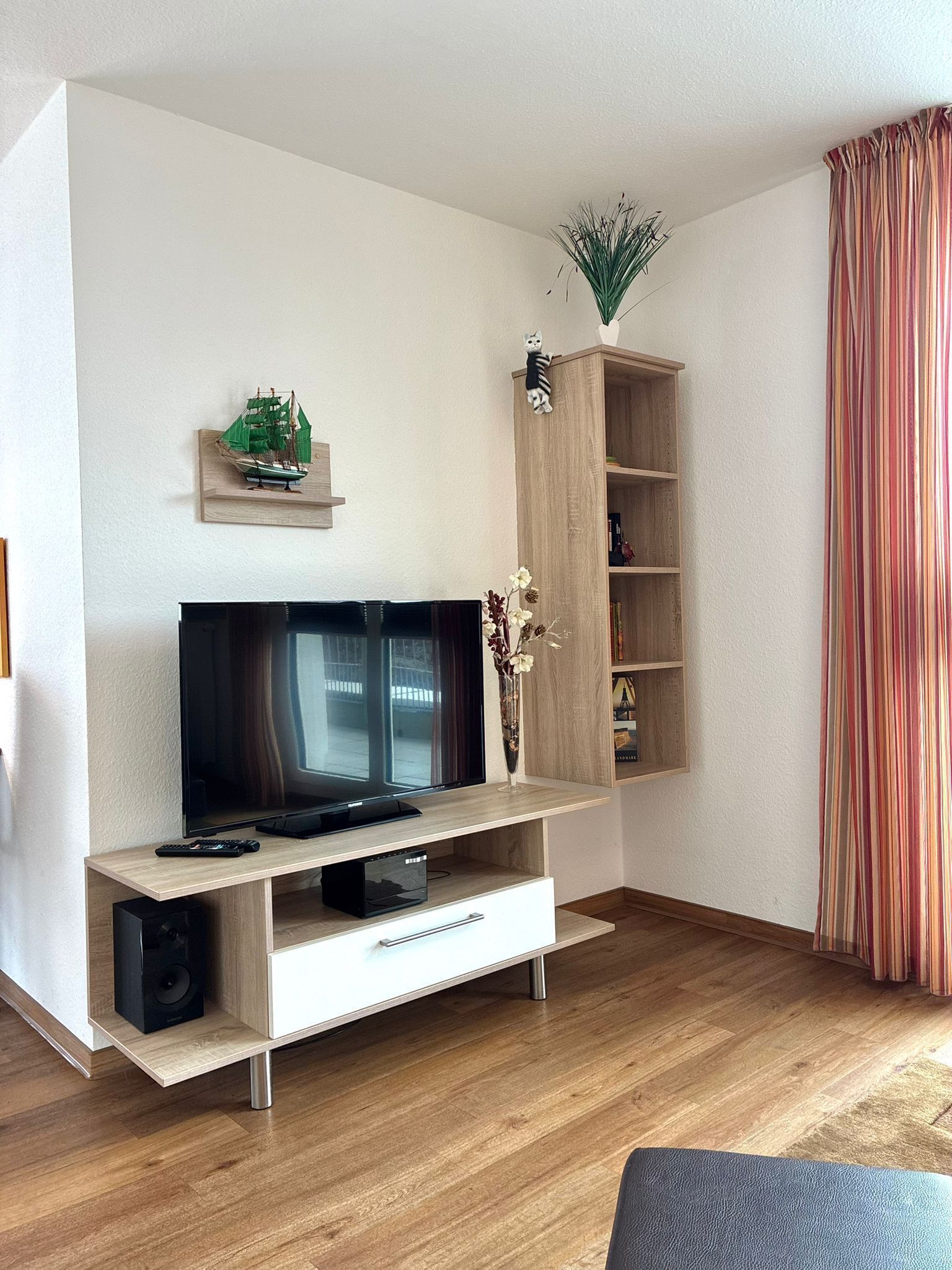 A modern living room with a flat-screen TV, wooden entertainment unit, and a small shelf with decorative items.