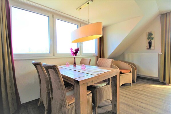 Un coin repas avec une table en bois et des chaises en osier, une lampe suspendue et de grandes fenêtres avec des rideaux.