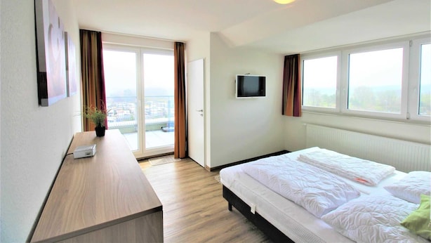 Habitación de hotel con una cama grande, un escritorio, un televisor y vistas a la ciudad a través de las ventanas.
