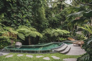 Un exuberante jardín con una piscina circular, terraza de madera y sillones de descanso.
