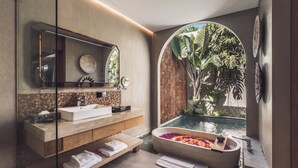 Un baño moderno con un espejo grande, dos lavamanos, una bañera llena de flores y vistas a una piscina y vegetación.