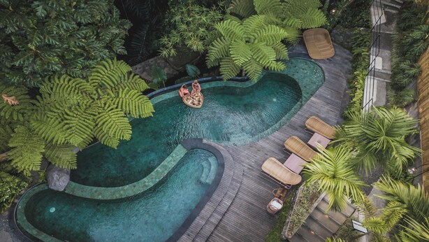 Vista aérea de una exuberante piscina infinita con terrazas rodeada de densa vegetación tropical, con solárium de madera y un pequeño bote.