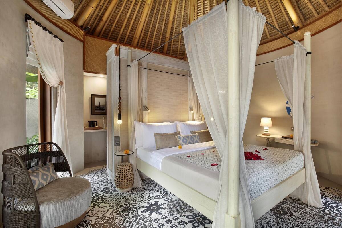 A four-poster bed with white bedding, a wicker chair, and patterned floor tiles.