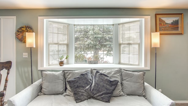 A light gray sofa with patterned pillows, a window with open shutters, and a framed painting on the wall.