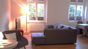 Sala de estar com sofá cinza, mesa redonda e luminária. Há janelas com persianas e um radiador.