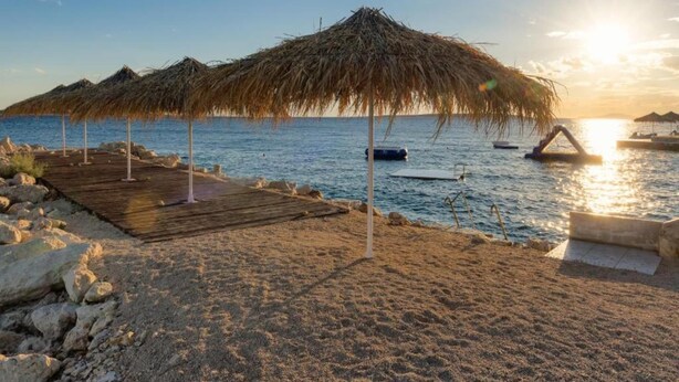 Une plage avec des parasols en chaume, une terrasse en bois et des bateaux sur l’eau.