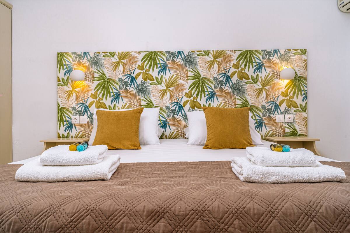 Ein Hotelzimmer mit einem Bett, zwei Nachttischen, einer Wanduhr und einem Kopfseitenverkleidung mit tropischem Blattmuster.