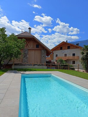 A swimming pool with a wooden deck, a stone building with a tiled roof, and a sign reading 'CorgiBauerhof'.