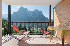 A balcony with two red chairs, a wooden table with glasses and a pitcher, and a mountain view.