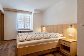 Una cama de madera con colchón blanco y ropa de cama floral, una mesita de noche con lámpara, y una ventana con cortinas traslúcidas.