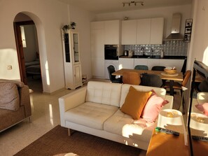 Ein Wohnzimmer mit einer Couch, einem Esstisch mit Stühlen, einer Küchenzeile mit Schränken und Geräten sowie einem Flur, der in einen weiteren Raum führt.