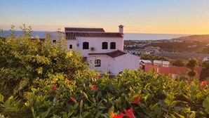 A white building with a tiled roof, surrounded by greenery and red flowers.