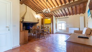 A spacious living area with a fireplace, wooden ceiling beams, a dining table with chairs, and a couch. - Cerboli Apartment with Sea View, Wi-Fi, Pool, and Air Conditioning (Riotorto)