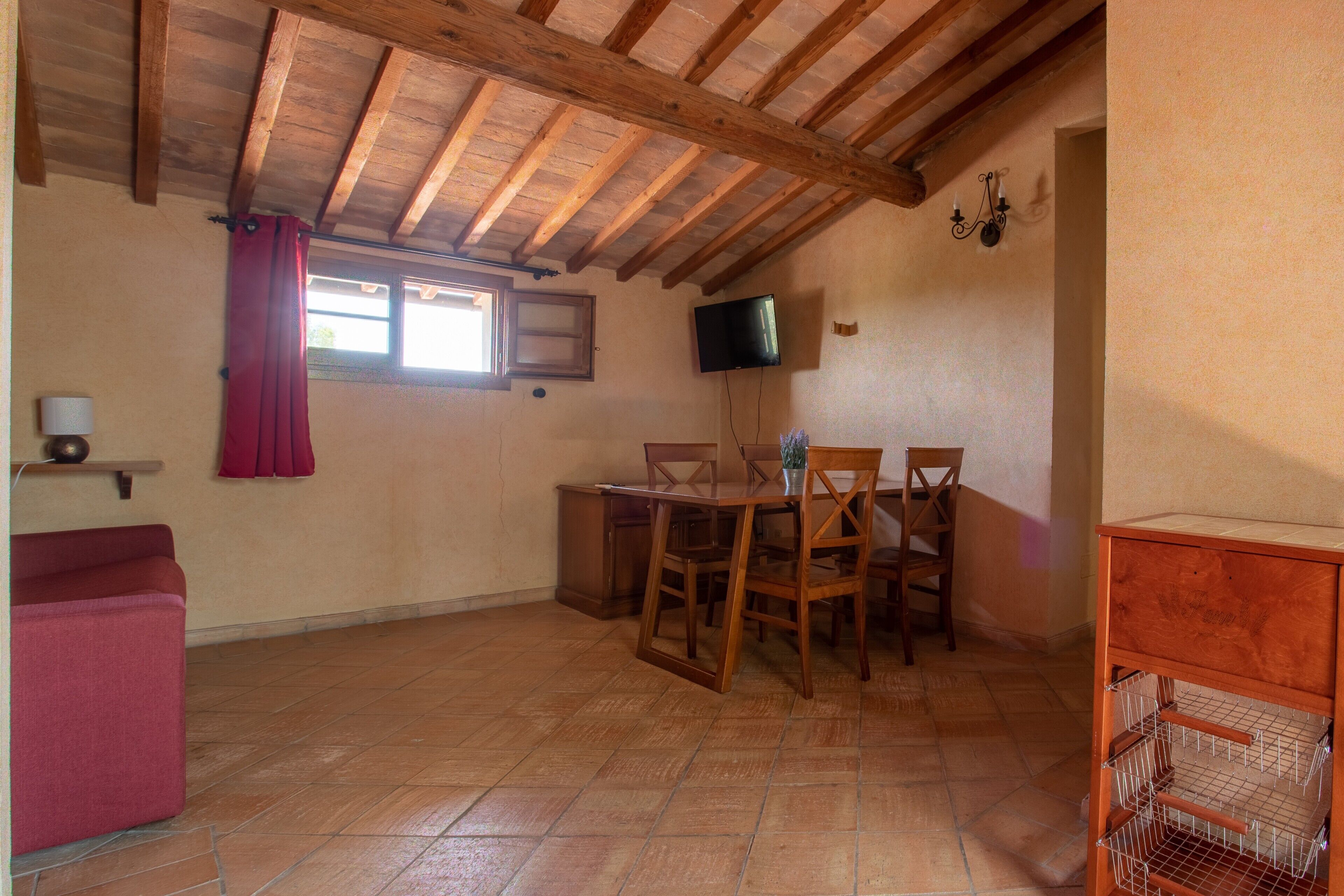 A dining area with a wooden table and chairs, a television mounted on the wall, and a red curtain.