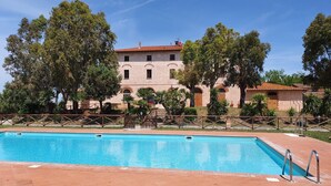 Ein Schwimmbecken mit Fliesenrand, umzäunt, im Hintergrund ein Gebäude mit Rundbogenfenstern und einem roten Ziegeldach.