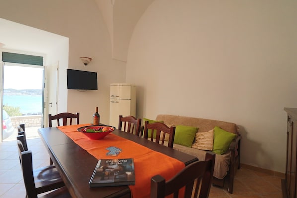 Un coin repas avec une table dressée pour deux personnes, un téléviseur fixé au mur et une vue sur l’extérieur par une porte ouverte.