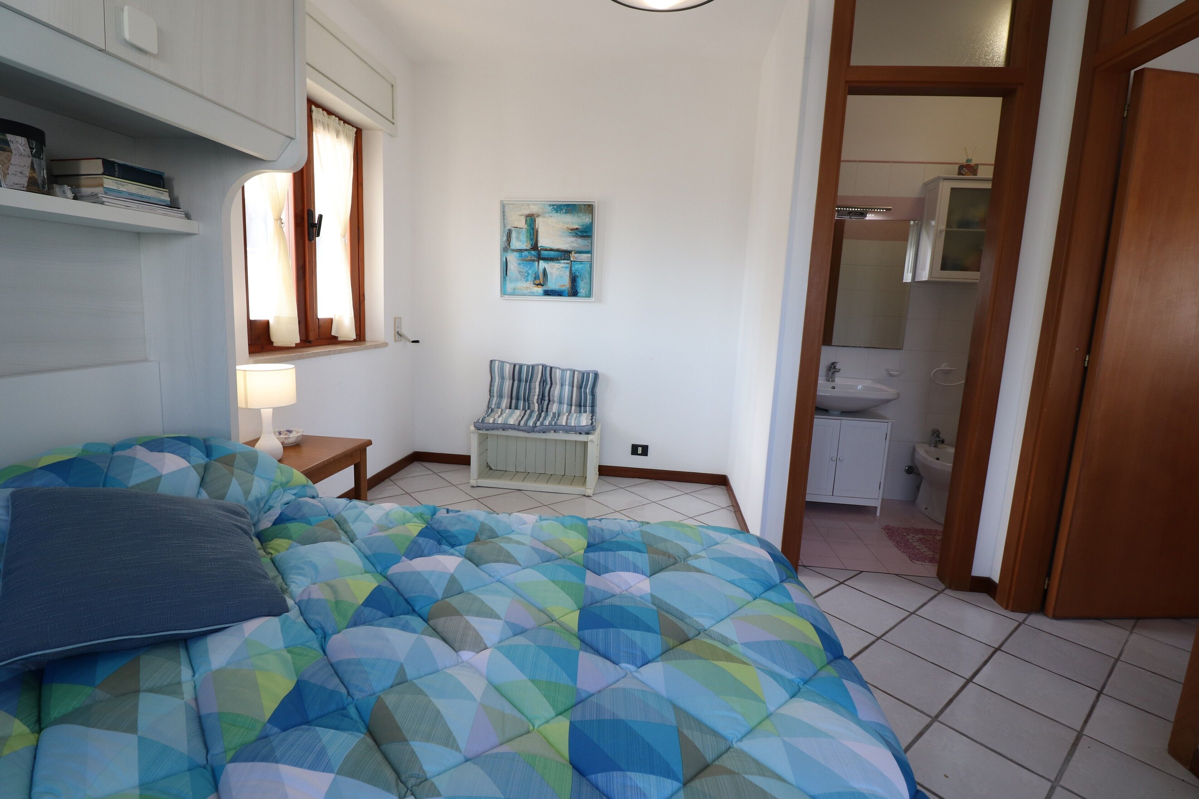 A bedroom with a bed, a bedside table, a lamp, a chair, a painting, and a bathroom visible through an open door.