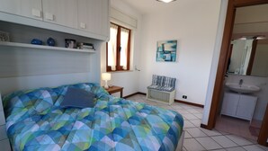 A bedroom with a bed, a nightstand, a chair, a painting, a bathroom with a sink, and a window with curtains.