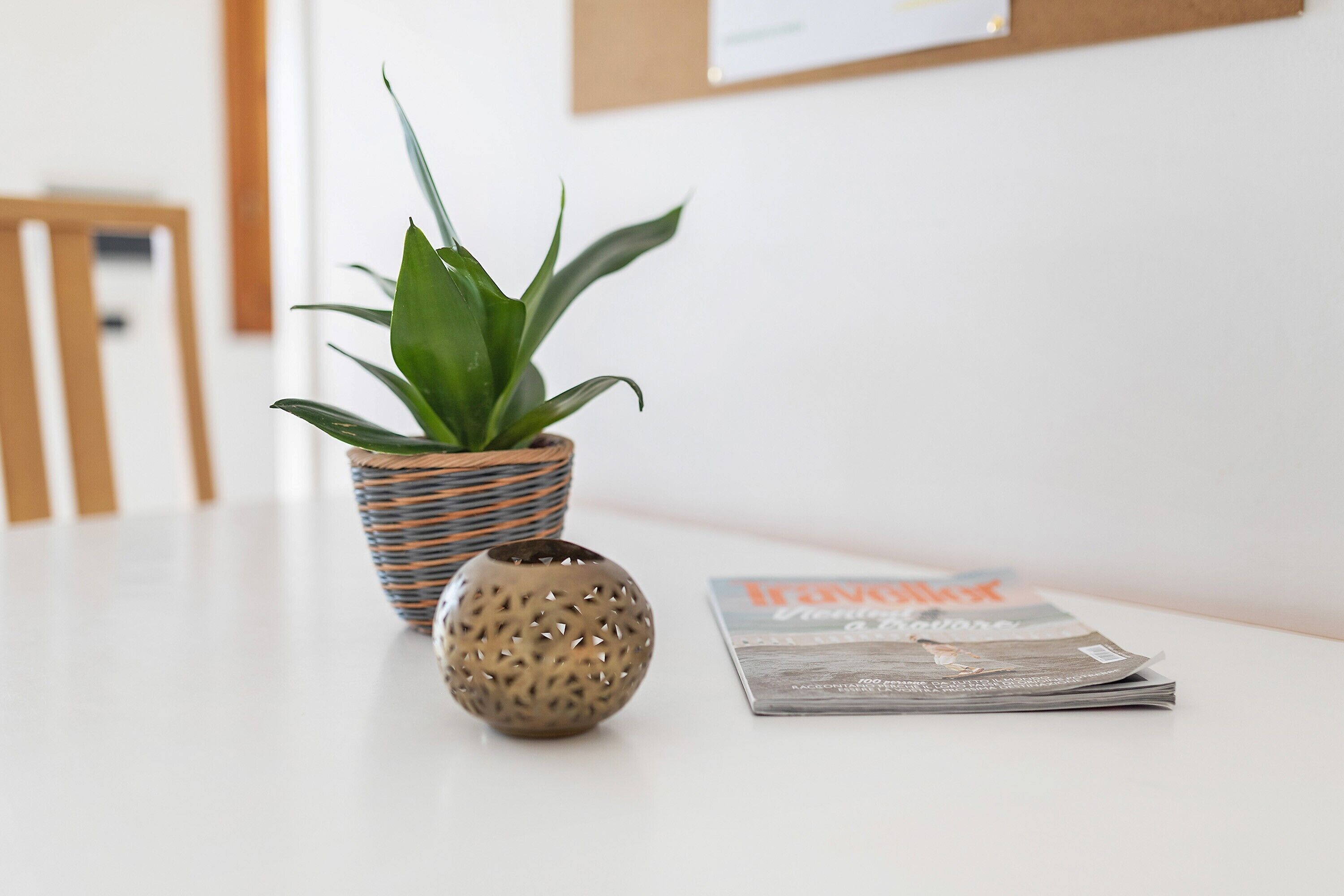 Een plant in een geweven pot, een decoratieve bol en een tijdschrift op een witte tafel.