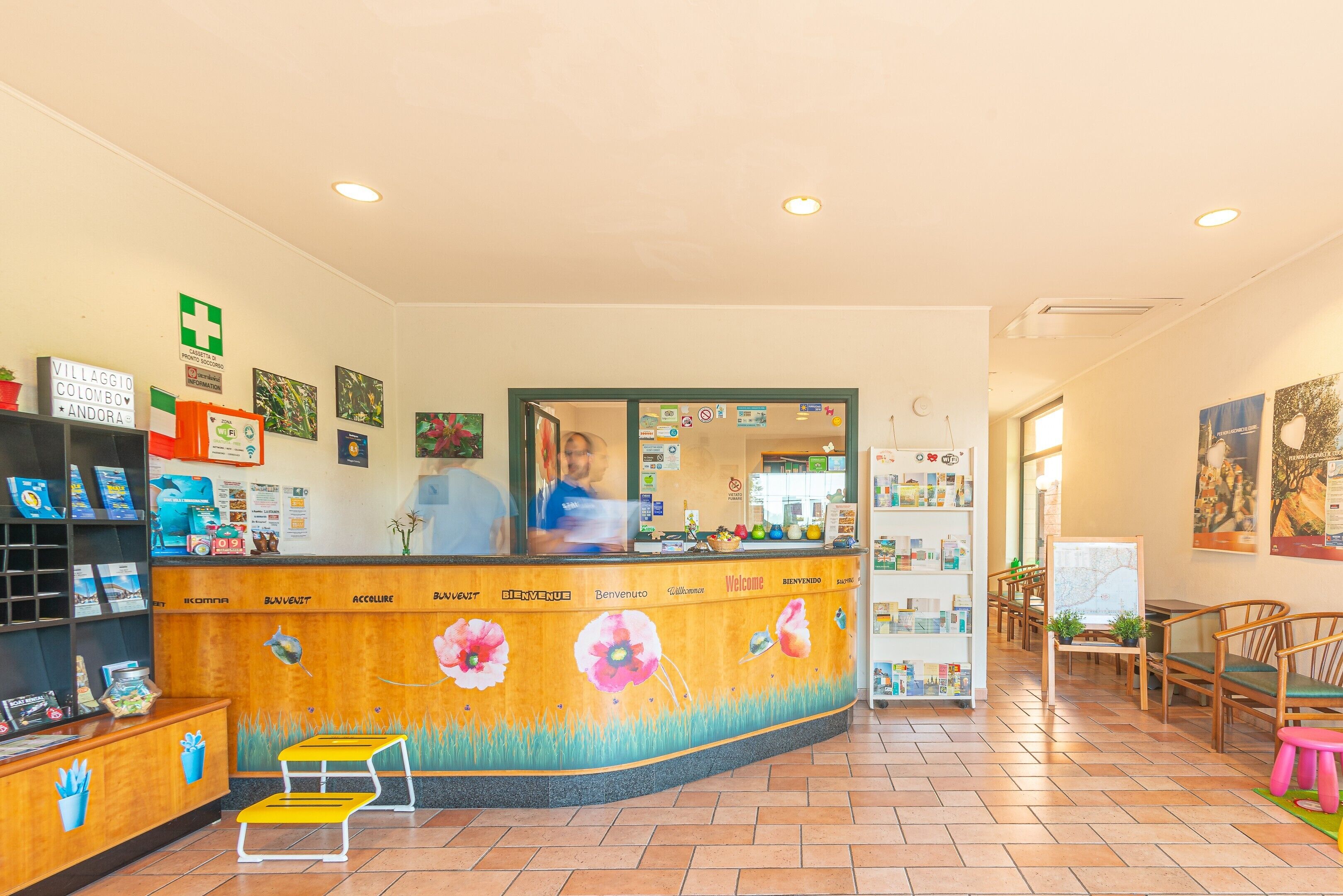Un comptoir d’accueil orné d’une fresque florale, un homme derrière le comptoir et un espace détente avec des chaises et un petit tabouret.