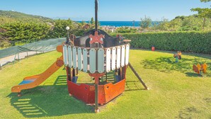 A playground with a slide and climbing structure, surrounded by grass and trees, with a view of the sea in the background. - New Management Holiday Home Villaggio Colombo V3 with Garden & Shared Pool (Andora (SV))