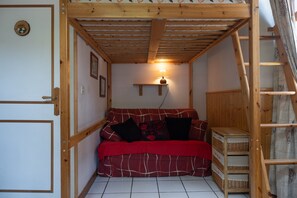 Une pièce chaleureuse avec un plafond en bois, un canapé rouge agrémenté de coussins noirs et rouges, une lampe fixée au mur et une échelle en bois.